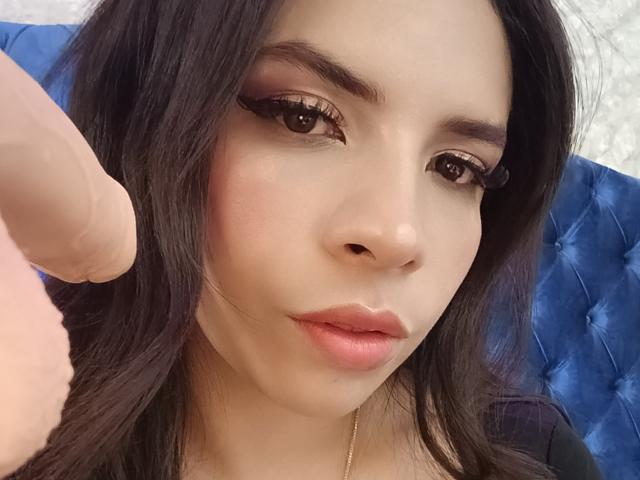 VioletaStone with long black hair, brown eyes, and pink lipstick, holding a realistic dildo in a selfie. Her expression is neutral, lips slightly parted. Blue tufted background.