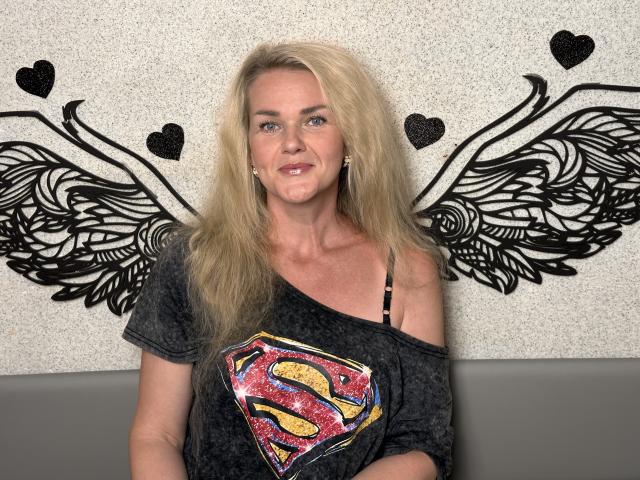PenelopaMurMiay with long blonde hair, grey off-shoulder Supergirl t-shirt, black bra strap visible, smiling, sitting, black wing tattoo behind her, grey bench.