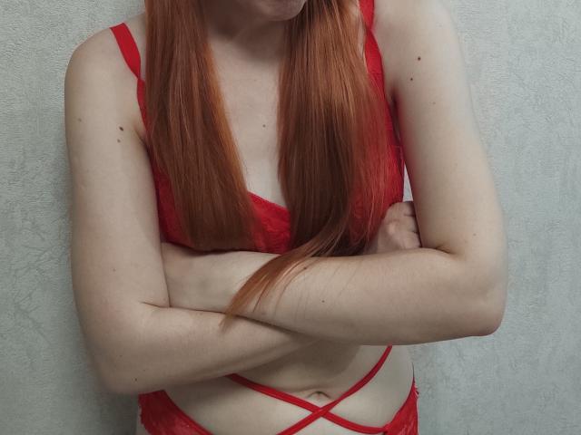 SheritaChieng with red lace lingerie, arms crossed, showing pale skin and long red hair, small tits, and a slim, flat-ass. She's standing against a plain wall.