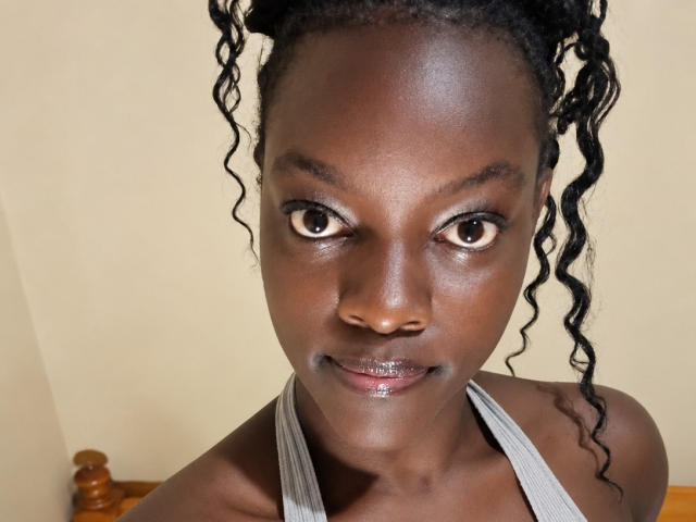 EvangelineDorrie has dark brown skin, curly black hair in puffs, brown eyes, and a shiny lip. She's wearing a grey halter top, showing her bare shoulders.