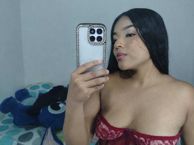 SheilaBrunette, con lunghi capelli neri, indossa un top rosso in pizzo senza spalline, tiene in mano un telefono con una custodia decorata con diamanti, sta scattando un selfie, mostrando il seno, in una camera da letto..