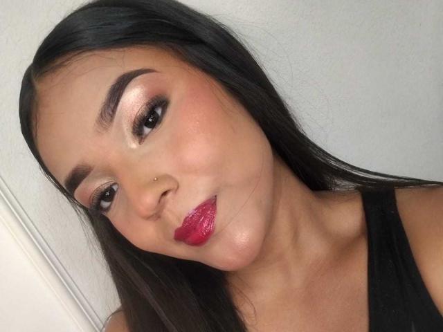 SheilaBrunette ha capelli castano scuro lucidi, sopracciglia folte, occhi rosa-marrone brillanti, rossetto rosso e un piccolo piercing al naso. Indossa una maglietta nera ed è sorridente..