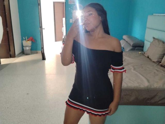 SheilaBrunette with long black hair, off-shoulder black dress with red, white, and blue stripes, holding phone in mirror, reflecting light, in bright blue room, standing by bed.