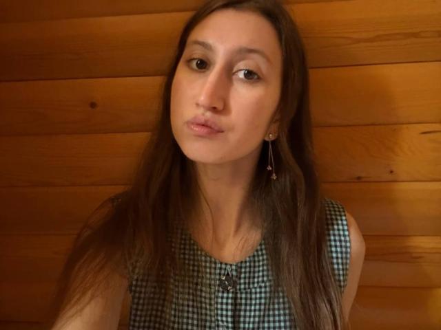 DorothaDemotta with long brown hair, pouty lips, and small earrings, wearing a green and white checkered sleeveless top, against a wooden wall. She's looking slightly to the side.