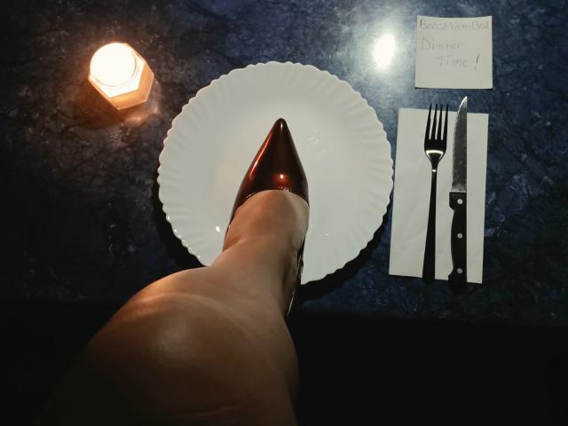 La jambe de BeccaVanBoa, avec des bottes marron brillantes pointant vers une assiette blanche, un couteau et une fourchette sur une table bleu foncé, avec une bougie allumée à gauche..