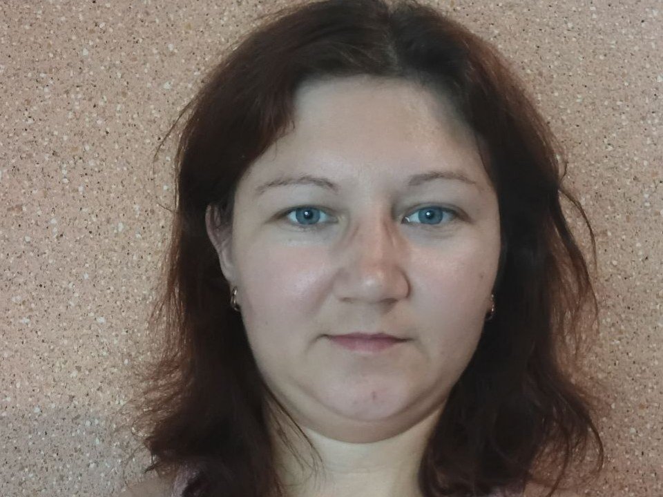OliviiaValter has pale skin, blue eyes, and brown wavy hair. She's wearing small earrings and has a slightly chubby face with a neutral expression.