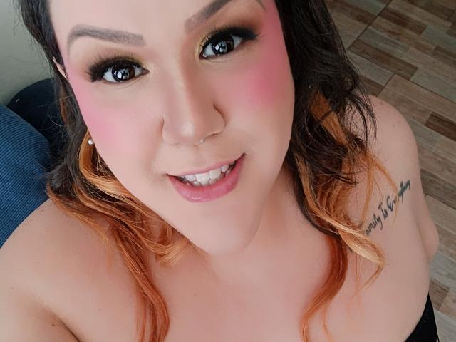 GiiannaMorrys with pink blush, nose ring, and smile, looking up with brown eyes, wavy blonde-to-brown hair, tattoo on right shoulder, and black strap top.