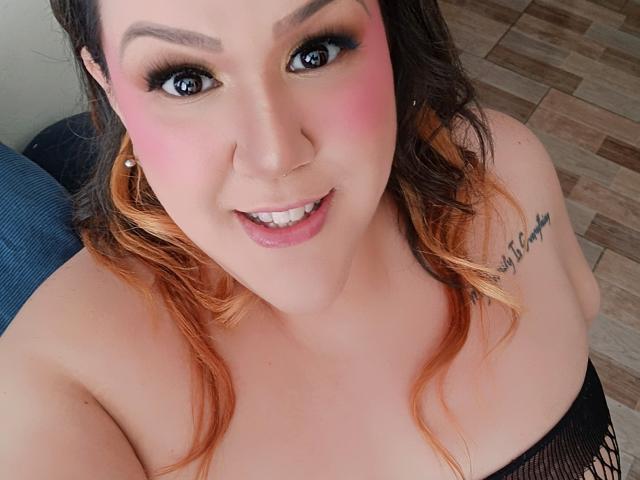 GiiannaMorrys has pink blush on her cheeks, black eyeliner, and orange-brown wavy hair. She's smiling with visible teeth, wearing a black top, and has a tattoo on her right shoulder.