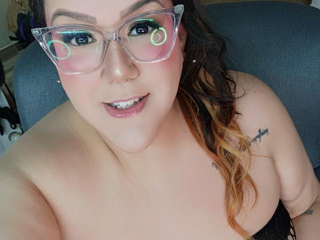 GiiannaMorrys with glasses, pink blush, nose piercing, smiling, wavy brown hair with orange tips, tattoos on shoulder, fair skin, black top, looking at camera, visible teeth.