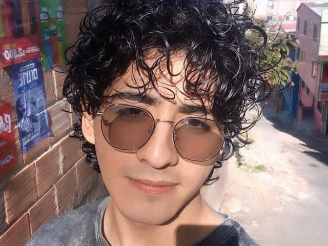 ElliotNoir has curly black hair, tan skin, and is wearing aviator sunglasses. He's sporting a grey shirt, and his slightly smiling face is in the sunlight.