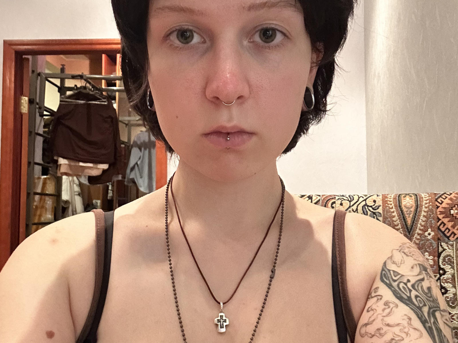 VasilikiAndersson has pale skin, short black hair, a nose ring, and a lip piercing. She wears a black tank top, necklace with a cross, and hoop earrings. Tattoo on right shoulder.