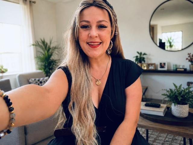 SamanthaGold, avec de longs cheveux blonds, une robe noire, souriante, tendant son bras pour un selfie, portant un collier, un bracelet et des boucles d'oreilles dorés, dans une pièce lumineuse. Ses seins sont visibles sous la robe..