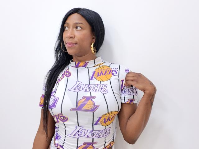 Sttefany, met donkere huid, lang zwart haar, draagt een witte LA Lakers shirt, gouden oorbellen, kijkt opzij, heeft haar hand op haar schouder, glimlacht licht en laat haar borsten zien..