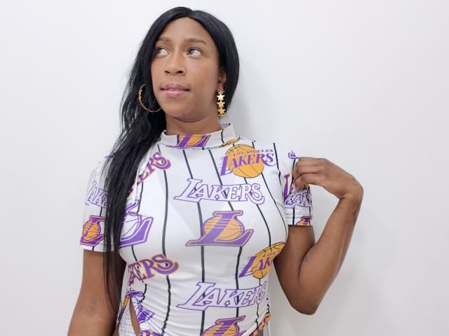 Sttefany, com cabelos longos e pretos, vestindo um vestido com tema do time Lakers, brincos de argola dourados e brincos dourados, está em pé em frente a uma parede branca, tocando o cabelo. Seus seios são de tamanho médio..