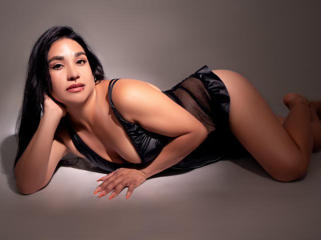 ElenaaParker with black hair, lying on her side, wearing a black mesh lingerie top, exposing her small tits and butt, looking at the camera, with orange nails.