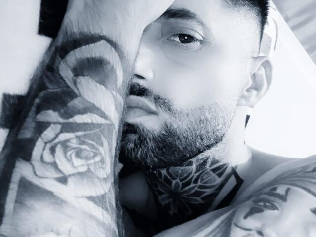 TommyDum, con barba, brazos tatuados y un tatuaje de rosa en su brazo derecho, mirando hacia arriba con una expresión seria, en un selfie en blanco y negro..
