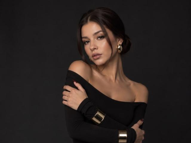 ChristianaKryzak with dark hair in a bun, wearing an off-shoulder black top, gold earrings, and gold cuff, arms crossed, looking seductively at the camera.