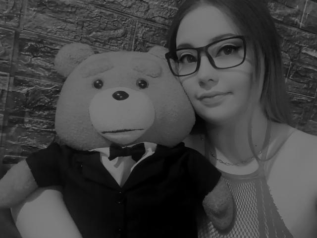 Black-and-white selfie of FoxyLyahot with glasses, wearing a mesh dress, and a large teddy bear in a suit, arms around each other, FoxyLyahot's tits slightly visible.