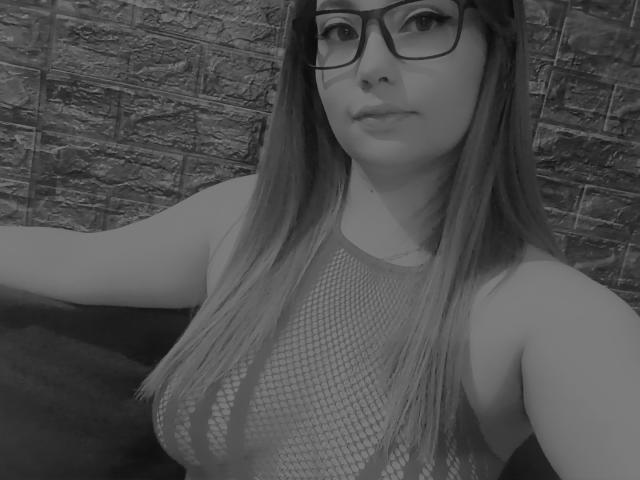 Grayscale selfie of FoxyLyahot with long hair, glasses, and a mesh top, showing her tits, sitting against a textured brick wall. FoxyLyahot's arm is resting on a dark surface.