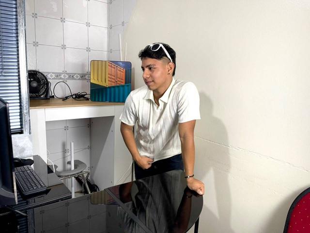 RichiiHott, standing, white short-sleeve shirt, black skirt, black pants, sunglasses on head, right hand on skirt, left hand on desk, white tiled wall, desk with laptop, chair.