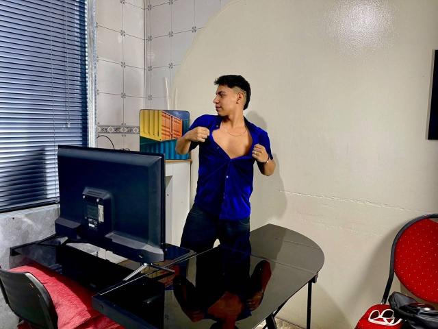RichiiHott, standing in a room, wearing an unbuttoned blue shirt, revealing a toned chest, with black hair, near a black desk and computer, looking to the side.