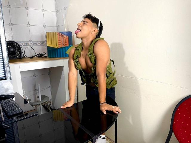 RichiiHott with short black hair, green sleeveless top, and exposed tits, leans over black table, tongue out, hands on table, wearing black pants, in a brightly lit room.