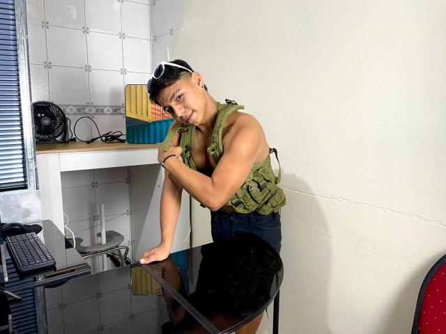 RichiiHott leaning on a glass table, wearing a green military-style vest, black shorts, and white sunglasses, with short black hair, smiling, showing a toned, muscular, tan-skinned body.