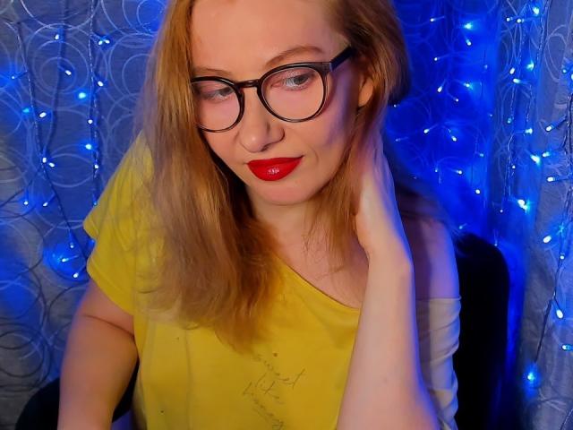 PaulinaSunny with red lipstick, glasses, and yellow shirt, hand in hair, blue fairy lights in background, smiling, fair skin, red hair, medium tits.