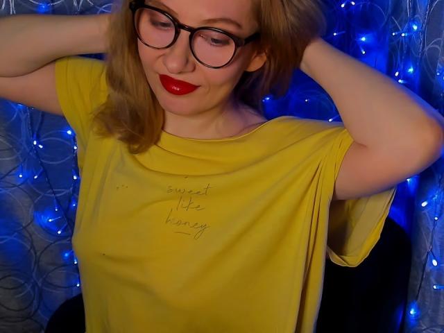 Paulina, lunettes, rouge à lèvres rouge, chemise jaune, mains dans les cheveux, guirlande lumineuse bleue, et rouge à lèvres rouge, regardant vers le bas, souriant. Ses seins ne sont pas visibles..