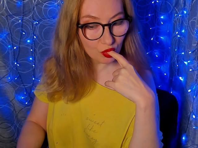 PaulinaSunny with red lipstick, black glasses, yellow shirt, biting her finger, blonde hair, blue fairy lights in background, looking seductive. Her tits are slightly visible under the shirt.