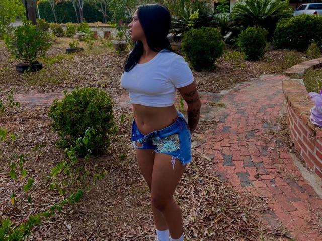 AlanaLuxe stands in a yard, wearing a white crop top, blue frayed shorts, white socks, and has black hair, tattooed arms, and a tan complexion, looking to the side.