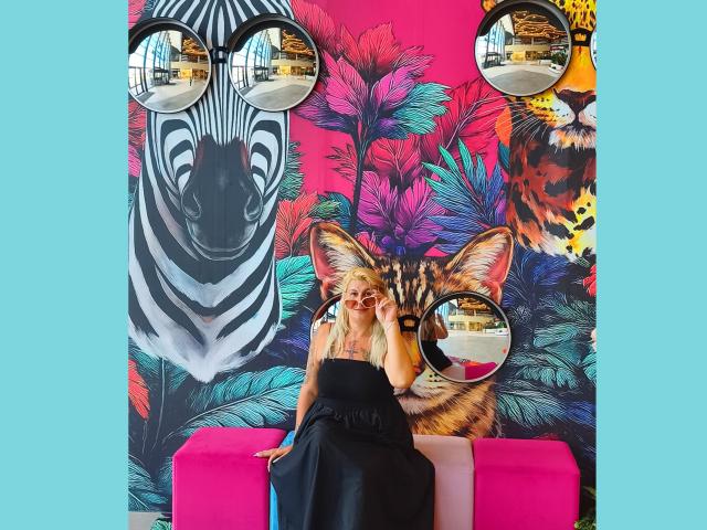 LilithGreenn è seduta su una panchina rosa, indossa un abito nero, e sullo sfondo colorato del muro si vedono i volti di una zebra e di un tigre, riflessi in due specchi rotondi..