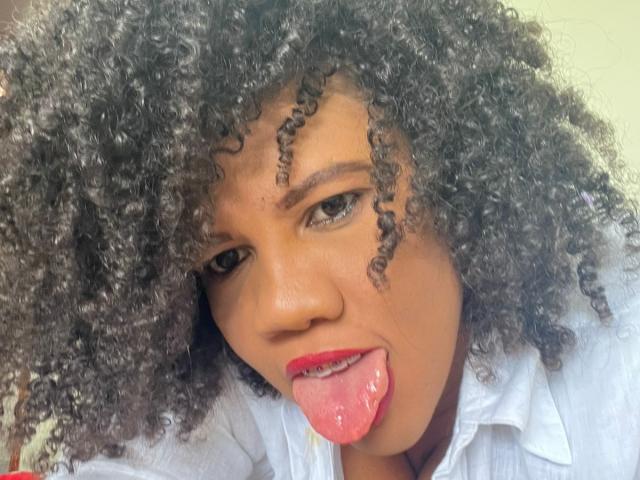 PamelaHorny69, avec ses cheveux noirs bouclés, sa langue rose en dehors, portant une chemise blanche. Son rouge à lèvres rouge est visible. Elle a un teint brun clair et regarde vers le haut..