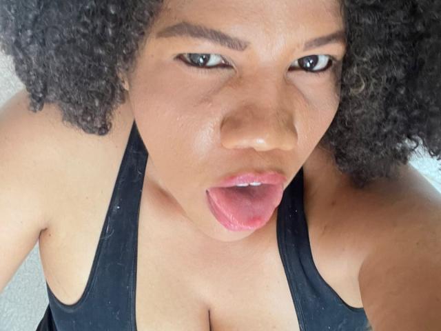 PamelaHorny69 heeft krullend zwart haar, donkerbruine ogen en draagt een zwarte tanktop. Ze steekt haar roze tong uit met haar mond open. Haar borsten zijn zichtbaar..