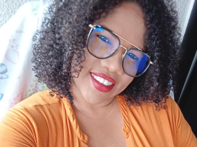PamelaHorny69 has curly black hair, wearing orange top, blue reflective sunglasses, and red lipstick, smiling at the camera with white teeth. Boobs not visible, pussy not visible.