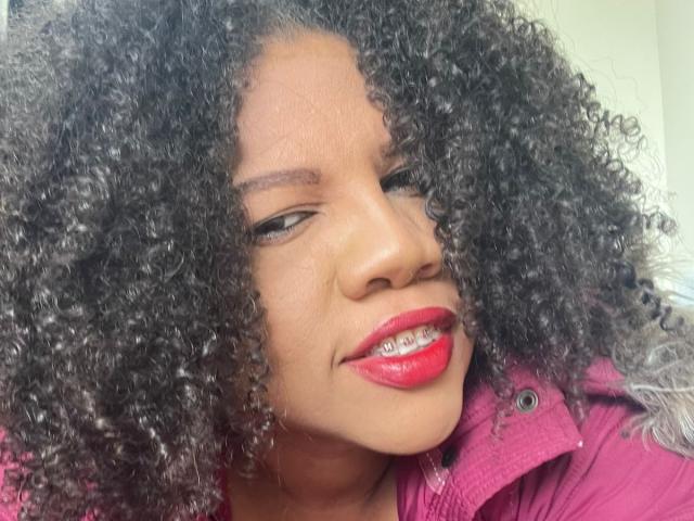 PamelaHorny69 with curly black hair, red lipstick, and a pink jacket, smiling slightly with her eyes half-closed. Her nose is centered, and her teeth are visible.