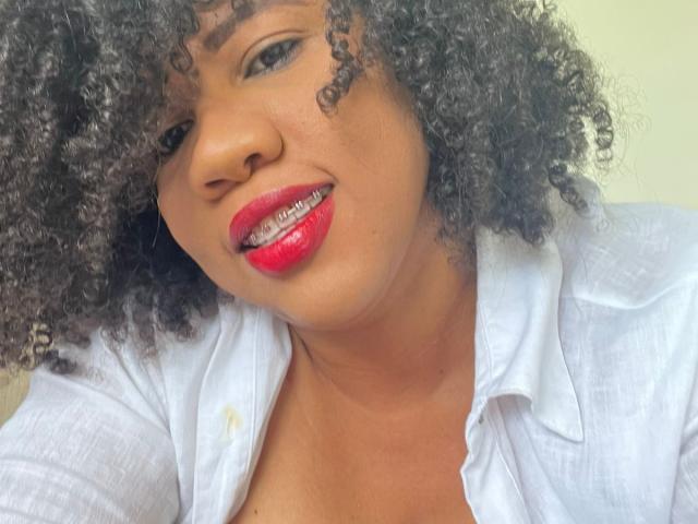 PamelaHorny69 with curly black hair, red lipstick, and white shirt, showing teeth in a slightly open-mouthed smile, has medium brown skin and a cute, sexy face.