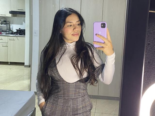 SophiaMarx with long black hair, wearing a checkered dress over a white turtleneck, taking a mirror selfie with a purple phone, showing a cute face and a small ass.