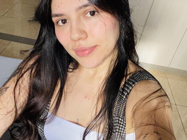 SophiaMarx with long black hair, light skin, small red blemishes on face, wearing a black and white checkered top, showing a small tattoo under her cleavage, smiling.
