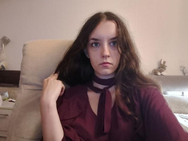 Fransisca Liversedge, con i capelli castano ondulati, gli occhi azzurri e un'espressione seria, indossa una camicia color borgogna con un fiocco, è seduta su una sedia beige e sta toccando i capelli..