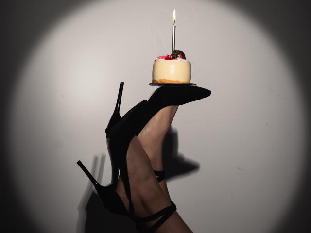 Le pied de MiaReed, chaussé de talons hauts noirs, tenant un petit gâteau d'anniversaire avec une bougie allumée, sur une surface plane, dans l'ombre, avec un effet de projecteur..