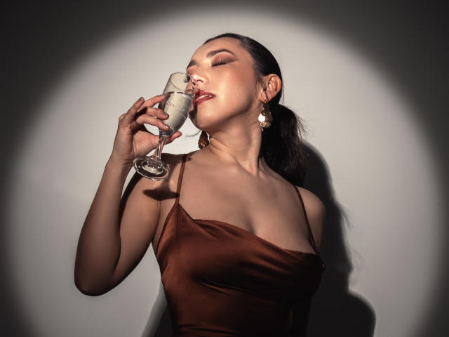 Mia Reed, les yeux fermés, sirotant un verre de champagne, vêtue d'une robe en satin marron, de boucles d'oreilles dorées, et ses cheveux noirs attachés en queue de cheval, sous un éclairage doux..