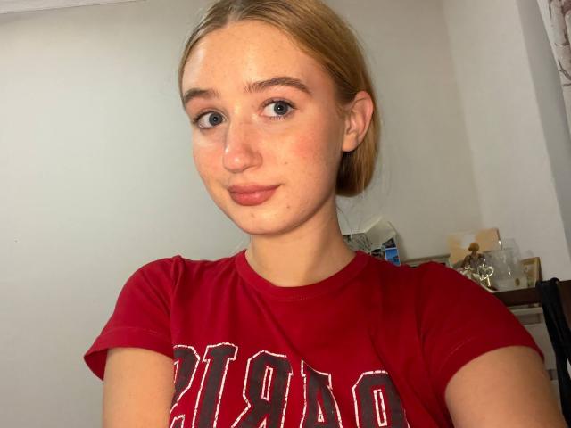 ShelliBetthauser with blonde hair, blue eyes, and freckles, wearing a red "DIAMOND" shirt, smiling subtly. Her small tits are slightly visible under the shirt.
