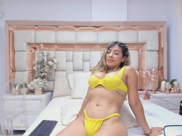 Kendra Miller, usando um conjunto de lingerie amarelo (sutiã e calcinha), sentada na cama, mostrando seus seios e vulva, cabelos castanhos longos, tatuagem no lado direito, sorriso confiante, ambiente de quarto..