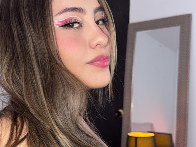 LorensDouce has pink eyeliner, glossy pink lips, and long brown hair, looking to the side with a slight smile. Her tits are partially visible, and she's wearing a black top.