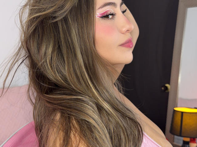 LorensDouce with pink eyeliner, pink blush, and pink lipstick, looking to the side with long brown hair, showing her tits slightly, in a room with a lamp.