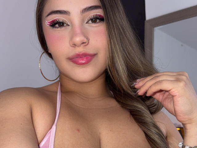 LorensDouce with pink eyeliner, glossy pink lips, hoop earrings, and long brown hair, touching her hair, showing her tits in a pink halter top.