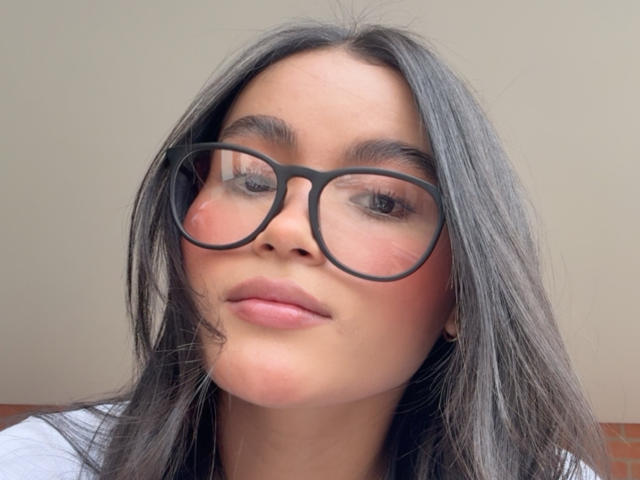 MiiaPalmer tiene gafas grandes y redondas, piel marrón media y cabello largo y oscuro. Lleva un rubor ligero en las mejillas y un leve, sensual bostezo..