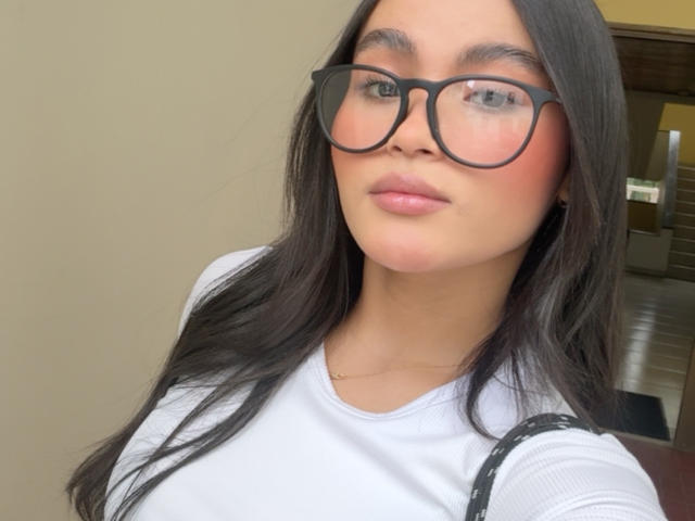MiiaPalmer has dark, wavy hair, glasses, and a serious expression. She's wearing a white shirt, showing her tits slightly, with a subtle pink blush on her cheeks.