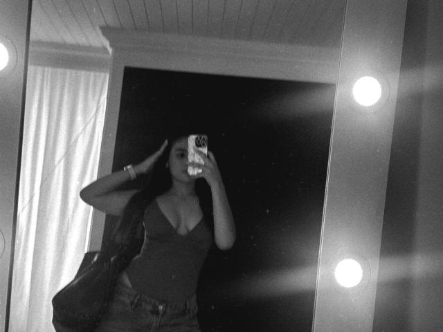Black-and-white selfie of MiiaPalmer with medium tits, wearing a low-cut tank top and jeans, holding a phone, standing in front of a mirror with lights.